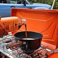 A person pouring traditional Indian chili paste into a black cooking pot on a portable camping stove, outdoors, with a vehicle in the background, showcasing Indian culinary essentials.