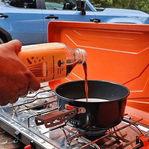 A person pouring traditional Indian chili paste into a black cooking pot on a portable camping stove, outdoors, with a vehicle in the background, showcasing Indian culinary essentials.